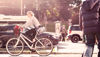 A female cyclist rides through town with cars, a bus and pedestrians in the background. 